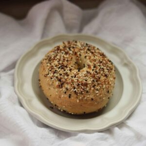 everything seasoned sourdough discard bagel on a small plate atop a white tea towel
