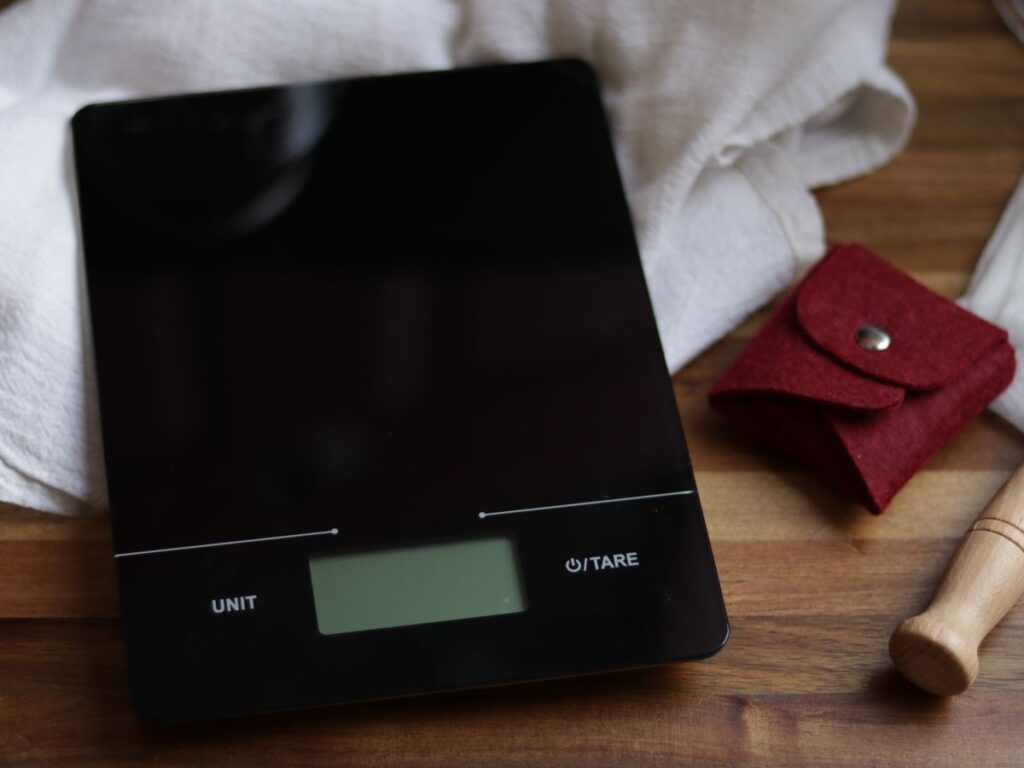 up close image of glass kitchen scale sitting on top of a wooden cutting board next to a white tea towel and small red pouch