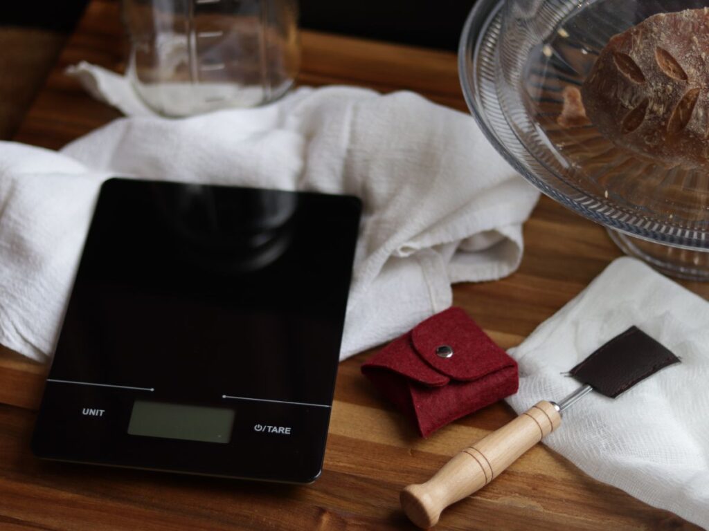 image of kitchen scale, sourdough scoring blades, a white tea towel, cheese cloth and a glass cake stand with sourdough bread in it sitting on a wooden cutting board